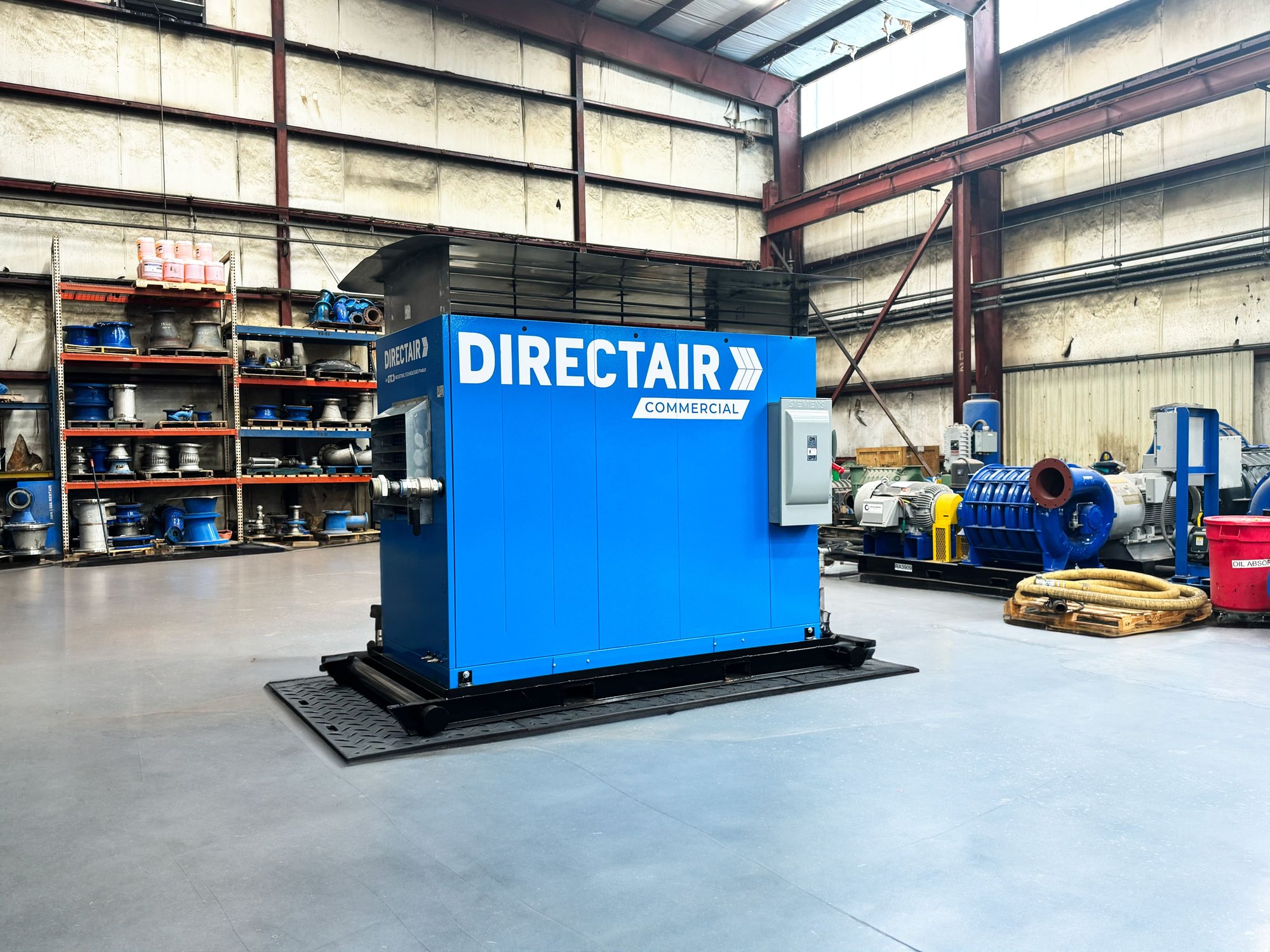 A large blue industrial compressor with the label "DIRECTAIR COMMERCIAL" in a warehouse with assorted mechanical equipment in the background.