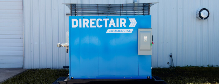 A blue industrial air compressor unit for DIRECTAIR Commercial against a metal building wall with a wall clock showing 10:10 to the right.