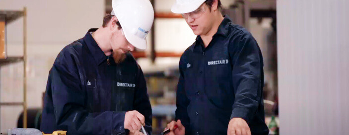 Two technicians in dark blue jackets with "DIRECTAIR" logos consulting over notes in an industrial workshop.