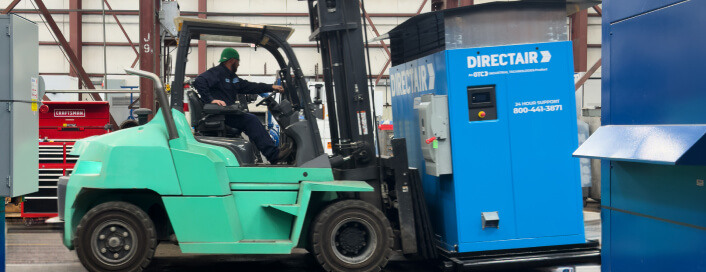 Worker in green helmet operates a teal forklift near a blue "DIRECTAIR" machine in an industrial setting.