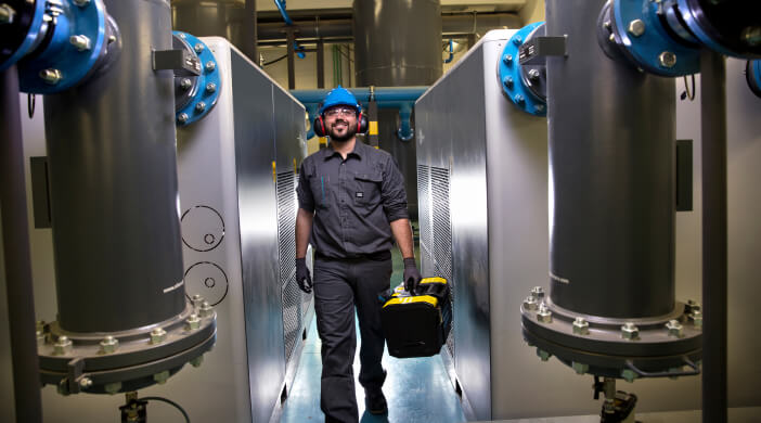 Man in an industrial facility carrying a toolbox, surrounded by large machinery.