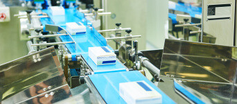 An industrial conveyor belt in a manufacturing facility carrying uniform white boxes with blue labels, with a selective focus and blurred background highlighting the machinery.