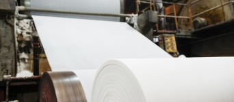 Close-up view of a large roll of white paper in an industrial setting with machinery in the background.